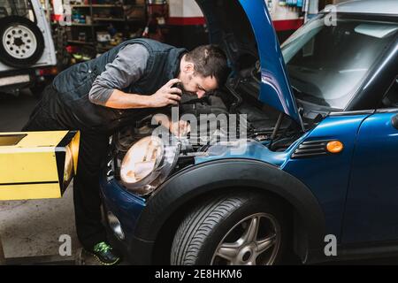 Junge fokussierte bärtige Wartung Mann Reparatur zeitgenössischen Auto gegen hell Licht in der Werkstatt Stockfoto