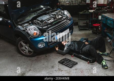 Junge fokussierte Auto-Service-Mann Überprüfung Fahrzeug mit offenen Motorhaube beim Liegen in der Werkstatt Stockfoto