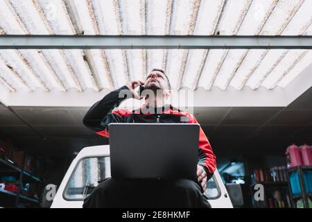 Von unten der männliche Meister mit Netbook reden auf Handy In der Nähe von Transport in der Garage Stockfoto