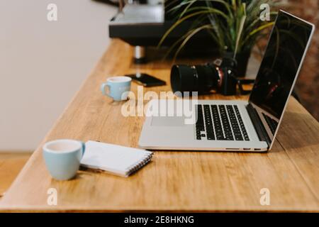 Arbeitsplatz des Freiberuflers mit modernen Netbook und professionelle Fotokamera Platziert auf Holztisch mit Tassen Espresso und Notizblock Im Café Stockfoto