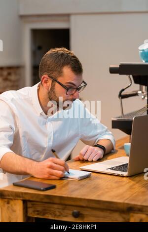 Fokussierter männlicher Barista, der an der Holztheke mit Laptop und sitzt Notizen im Notizbuch während der Arbeit im Café Stockfoto