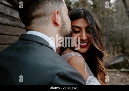 Seitenansicht des jungen Bräutigams in eleganten Anzug küssen lächelnd Ethnische Braut während in der Natur am Hochzeitstag stehen Stockfoto