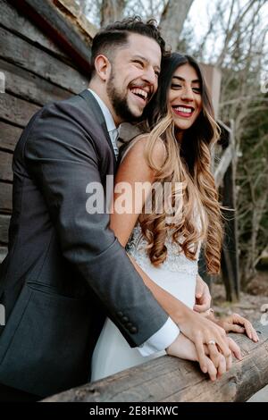 Seitenansicht des fröhlichen jungen bärtigen Bräutigams in Anzug lachend Und schmusen elegante ethnische Braut in weißem Kleid im Stehen Zusammen auf Holzbrücke n Stockfoto