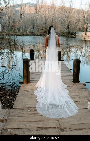 Rückansicht von unkenntlich wunderschöne junge Braut in elegantem Weiß Hochzeitskleid mit langen Schleier auf hölzernen Pier in der Nähe stehen see im Herbstwald Stockfoto
