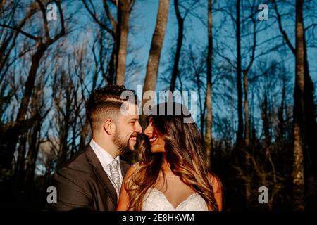Seitenansicht der jungen ethnischen Braut mit langen Haaren in Weißes Kleid lächelnd und umarmt Bräutigam mit geschlossenen Augen, während Zusammenstehend in der Nähe von Nadelbäumen Stockfoto