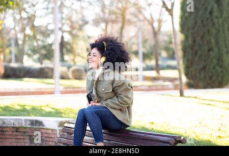 Aufgeregt junge afroamerikanische Frau Millennial mit lockigen Haaren in Stilvolles Outfit auf Bank im Park ruhen und hören Lieblings Titel in Kopfhörern Stockfoto