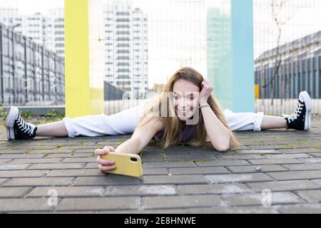 Ganzkörper glücklich fit weiblich tun Splits auf gepflasterten Gehweg Und Selfie auf dem Handy in moderner urbaner Umgebung Stockfoto