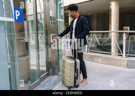 Seitenansicht des afroamerikanischen männlichen Touristen mit Koffer und In Schutzmaske drücken Taste des Aufzugs im Flughafen während Reisen während der Coronavi Stockfoto