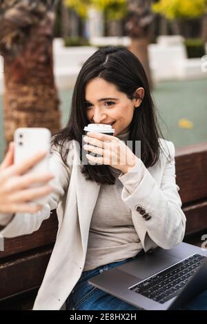 Entzückt junge Unternehmerin sitzt auf Bank mit Laptop und Selbstporträt mit Kaffee in Papiertasse zu gehen Während der Verwendung des Smartphones Stockfoto