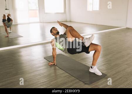 Seitenansicht des muskulösen jungen bärtigen männlichen Athleten liegend Matte in der Nähe von Spiegel und tun Glute Bridge März Übung während Allein trainieren bei viel Licht Stockfoto