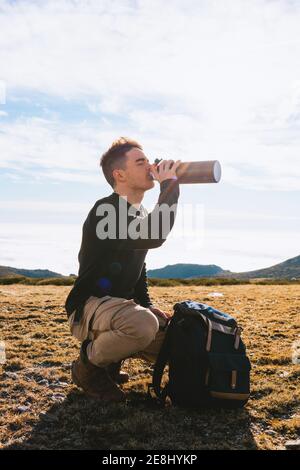 Seitenansicht des jungen stilvollen männlichen Reisenden in lässigem Outfit Sitzen auf Haunches in der Nähe von Rucksack und trinken heiße Getränke aus Thermoskannen während der Wanderung Stockfoto