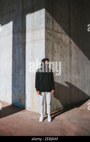 Trendy Kerl im Gesicht Maske wegschauen, während auf stehen Straße in der Nähe der schäbigen Mauer an sonnigen Tagen Stockfoto