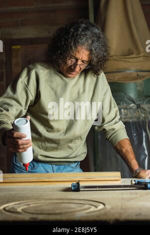Fokussierter reifer männlicher Zimmermann, der Klebstoff auf ein Stück Holz aufgibt Während der Tischarbeit in der Schreinerei Stockfoto