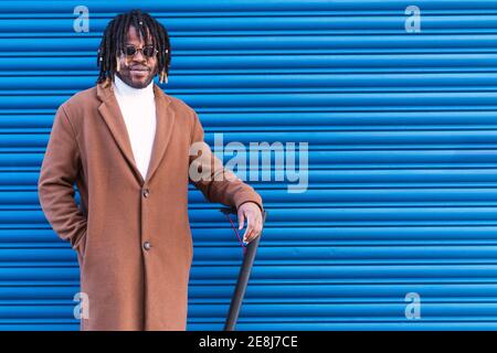 Selbstbewusster afroamerikanischer Hipster-Mann mit Afro-Zöpfen Stilvoller Mantel und Sonnenbrille stehen in der Nähe Elektroroller gegen blau Gestreift Stockfoto
