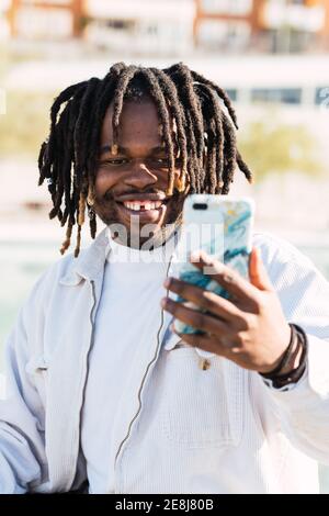 Stilvoller afroamerikanischer Rüde mit Dreadlocks und im trendigen Outfit Selbstporträt auf dem Smartphone in der Stadt an sonnigen Tag Stockfoto