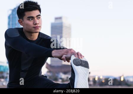 Fokussierter junger asiatischer Mann in Sportbekleidung, der Beine nahe Metall streckt Geländer beim Aufwärmen während des Outdoor-Fitnesstrainings bei Sonnenuntergang Stockfoto