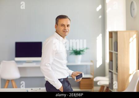 Porträt eines Geschäftsmannes mit einem Telefon in den Händen, das im Büro steht und sich auf den Tisch lehnt. Stockfoto