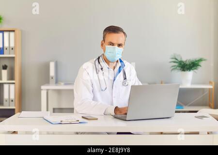 Seriöse Arzt in einer Schutzmaske sitzt an seinem Schreibtisch mit einem Laptop und Blick auf die Kamera. Stockfoto