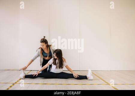 Lächelnd weibliche Trainer in Sportbekleidung helfen fröhliche Mädchen mit Stretching Beine beim Aufwärmen vor dem Fitnesstraining Stockfoto