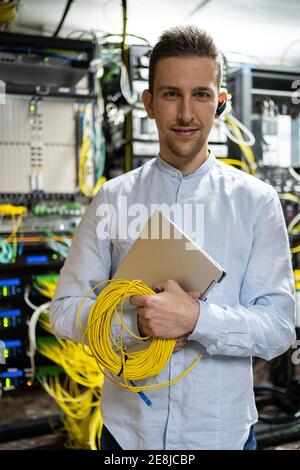 Positiver Mann im schnurlosen Headset, der mit Netbook und Kabeln steht Im Serverraum Stockfoto