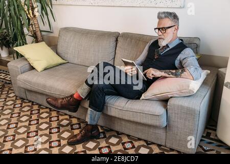 Trendy mittleren Alters Geschäftsmann Surfen im Internet auf Tablet während sitzen Mit gekreuzten Beinen auf Couch im Haus Stockfoto