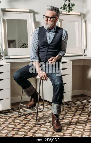 Trendy reifen Hipster Geschäftsmann in Brillen und trendige Kleidung suchen Weg auf Stuhl im Haus Stockfoto