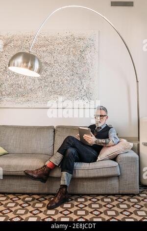 Trendy mittleren Alters Geschäftsmann Surfen im Internet auf Tablet während sitzen Mit gekreuzten Beinen auf Couch im Haus Stockfoto