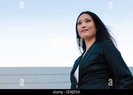 Von unten selbstbewusste asiatische weibliche Unternehmer zu Fuß entlang der Straße In der Nähe von Stadtgebäude und Blick weg Stockfoto