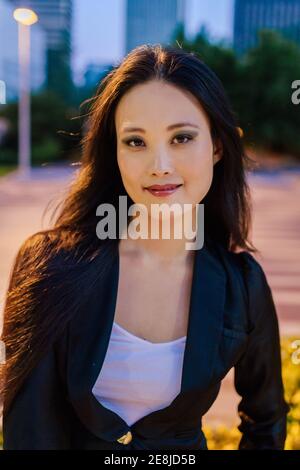 Fröhliche asiatische weibliche Unternehmer in smart casual Outfit Blick auf Kamera auf dem Hintergrund der Stadt am Abend Stockfoto