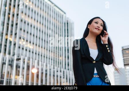 Niedriger Winkel der asiatischen weiblichen Unternehmer stehen in der Straße von Megalopolis und sprechen auf Handy, während geschäftliche Fragen zu diskutieren Und schauen awa Stockfoto