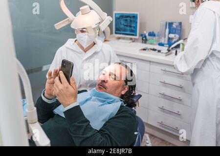 Patient mit Smartphone in der Zahnarztpraxis. Zwei Ärztinnen Vorbereitung des Verfahrens. Konzept der Zahngesundheit und Hygiene Stockfoto