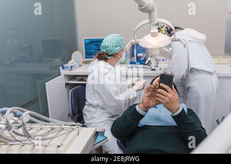 Patient mit Smartphone in der Zahnarztpraxis. Zwei Ärztinnen Vorbereitung des Verfahrens. Konzept der Zahngesundheit und Hygiene Stockfoto