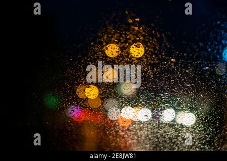 Regen fällt auf die Windschutzscheibe des Autos mit bunten Licht Bokeh am Abend während der Regenzeit. Stockfoto