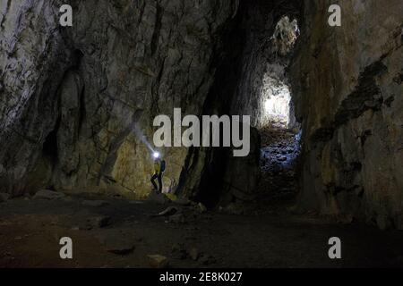 Frau mit Stirnlampe, die auf einem großen Felsen in einer Höhle steht, Licht, das vom Eingang der Höhle kommt Stockfoto