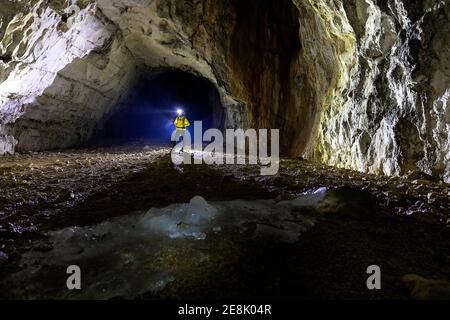 Frau in gelber Jacke in einer Höhle stehend, beleuchtet BU eine Hintergrundbeleuchtung, geschmolzene Stalagmiten aus Eis in der Front, Slowenien Stockfoto