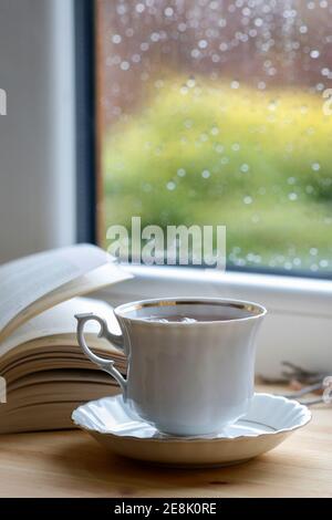 Porzellanbecher mit heißem Tee, weicher Decke und offenem Buch am Fenster. Entspannen Sie sich Stockfoto