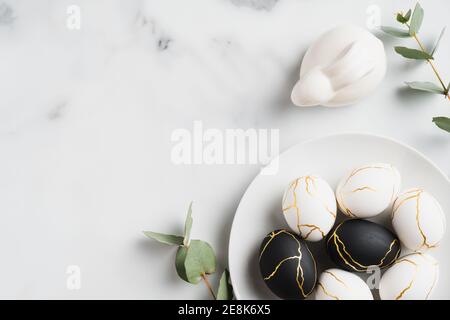 Elegante Ostern flach Lay Komposition. Schwarze und goldene Ostereier, Kaninchenhase, Eukalyptusblätter auf Marmorboden. Draufsicht mit Kopierbereich. Hap Stockfoto
