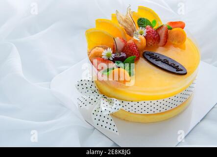 Orangenkuchen mit glücklichen Geburtstag und gekrönt mit Orange, Erdbeere, Heidelbeere und Cape Gooseberry auf weißem Stoffhintergrund, Kopierraum und Dessert c Stockfoto