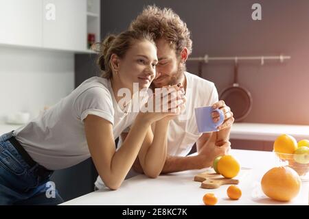Morgenkaffee in einem neuen Haus sanft küssen Frau junger Mann, die Frühstück in einer neuen modernen Küche und lächeln sich gegenseitig. Hübsches Mädchen füttert oder Stockfoto