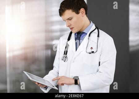 Ein junger, freundlicher Arzt überprüft einige Informationen auf seinem Computer-Tablet. Porträt eines Facharztes bei der Arbeit in der sonnigen Klinik Stockfoto