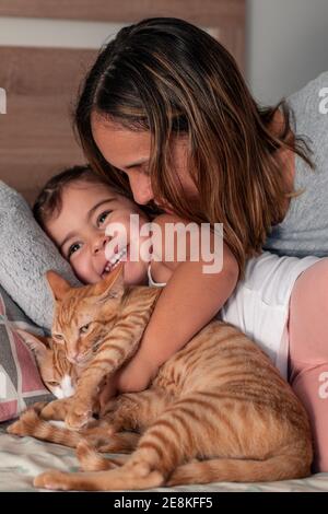 Kaukasische Mutter und Tochter, die mit ihrer kleinen Tabby-Katze auf dem Bett im Schlafzimmer spielen. Kitzelt sich gegenseitig und lacht fröhlich. Stockfoto