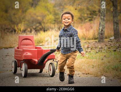 Fröhlicher und lächelnder Junge, der an einem Herbstnachmittag im Park in Bramble, Virginia, läuft und seinen roten Wagen zieht. Stockfoto