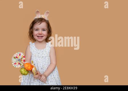 Emotionales, positives Porträt eines kleinen Mädchens mit Hasenohren auf dem Kopf, das einen Osterstrauß aus Eiern, Hühnern hält. Isolierte hellorange Hintergrundfarbe Stockfoto