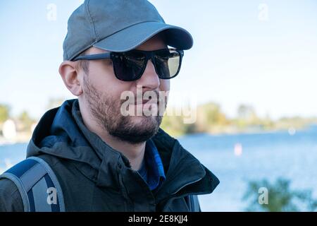 Bärtiger Mann in Sonnenbrille und Baseballmütze vor der Kulisse eines Sees oder Flusses an einem sonnigen Herbsttag. Konzept der Erholung im Freien, Einsamkeit oder Stockfoto