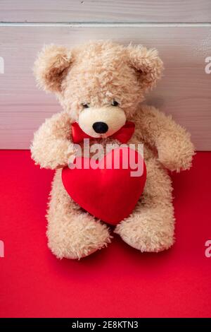 Valentinstag. Teddybär, Single, allein hält ein großes rotes Herz sitzt auf rotem Boden, Holzwand Hintergrund. Stockfoto