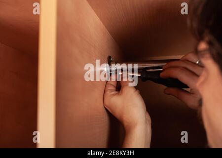 Ein Mann dreht ein Rohr für einen Aufhänger in einem Schrank mit einem Schraubendreher. Die Querlatte zum Aufhängen der Kleidung. Stockfoto