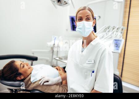 Porträt einer Zahnärztin in ihrer Klinik. Selbstbewusste Frau Zahnarzt trägt schützende Gesichtsmaske Blick auf die Kamera mit einem weiblichen Patienten in Delle Stockfoto