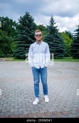 Ein hübscher junger Mann mit europäischem Aussehen trägt eine Sonnenbrille und ist mit einem Hemd und einer Jeans bekleidet. Der Kerl geht die Straße hinunter, er ist stilvoll gekleidet Stockfoto