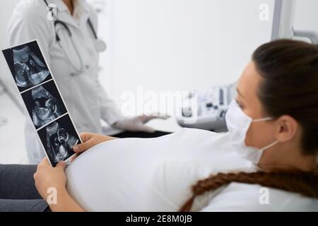 Glückliche Frau in Erwartung auf medizinische Couch liegen und in den Händen halten Bilder nach Ultraschall-Screening. Arzt und Patient tragen Gesichtsmasken während Stockfoto