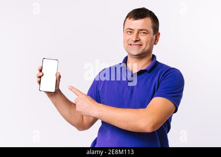 Kaukasischer Mann in blauem T-Shirt sieht glücklich an der Kamera und zeigt seinen Zeigefinger an den Mocks up Smartphone-Bildschirm. Grauer Hintergrund. Stockfoto